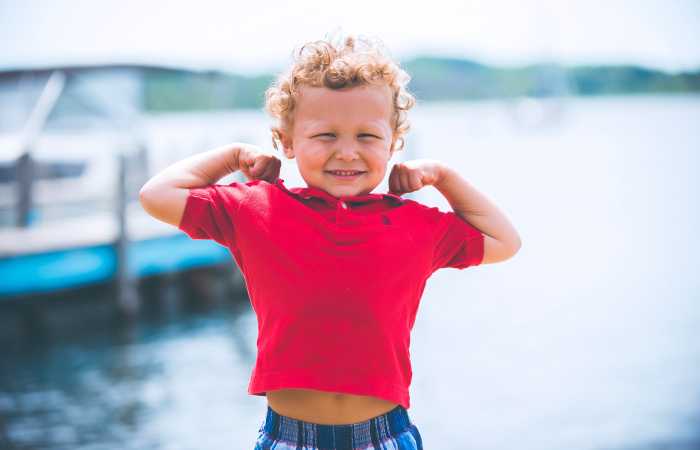 Curly haired young boy in a red polo flexes his arms in a strong-man pose and smiles by a lake