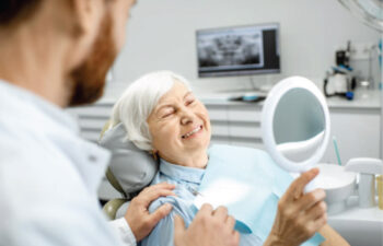 senior woman looks in a handheld mirror at her smile as the dentist looks on